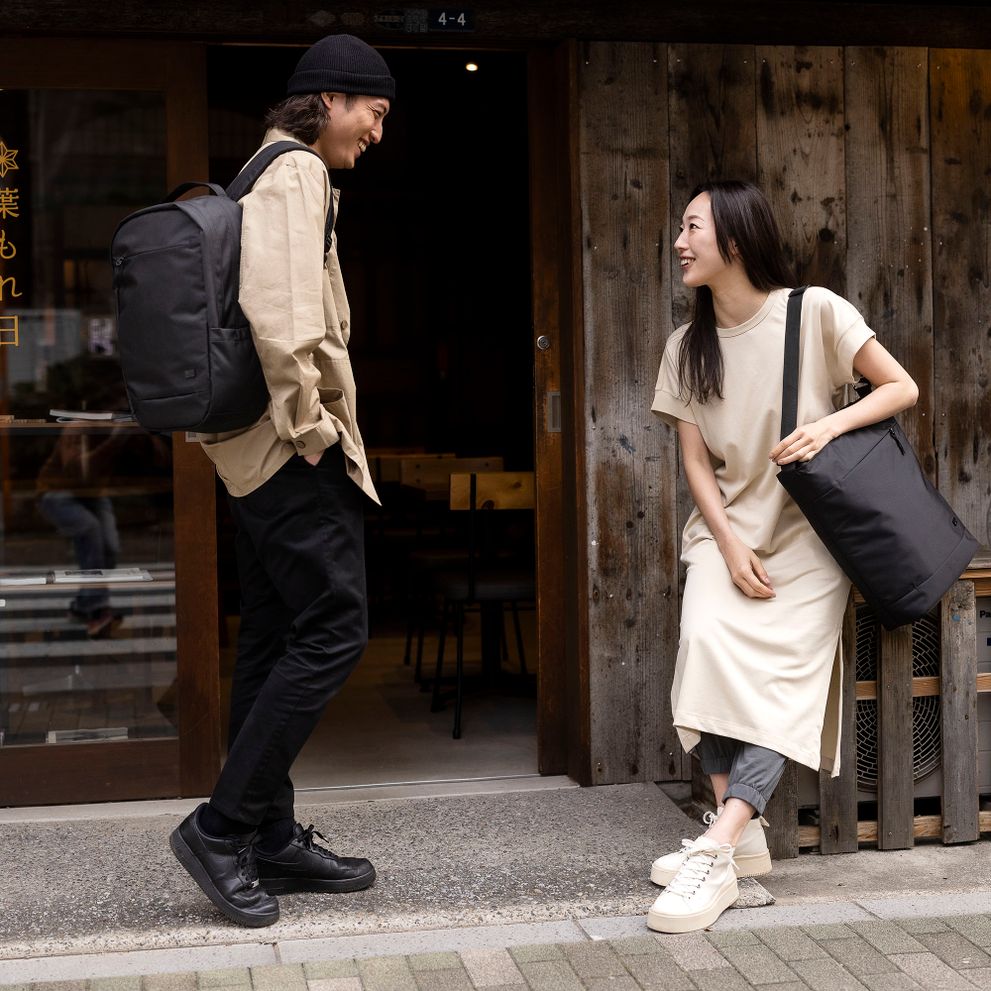 A couple laugh outside of a shop, holding black Case Logic Invigo tote and Case Logic Invigo backpack.