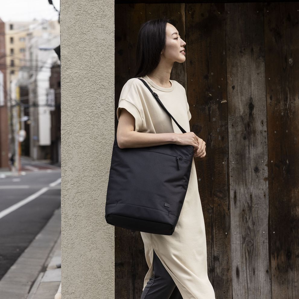 A woman leans on the wall by a city street, carrying a black Case Logic Invigo tote.