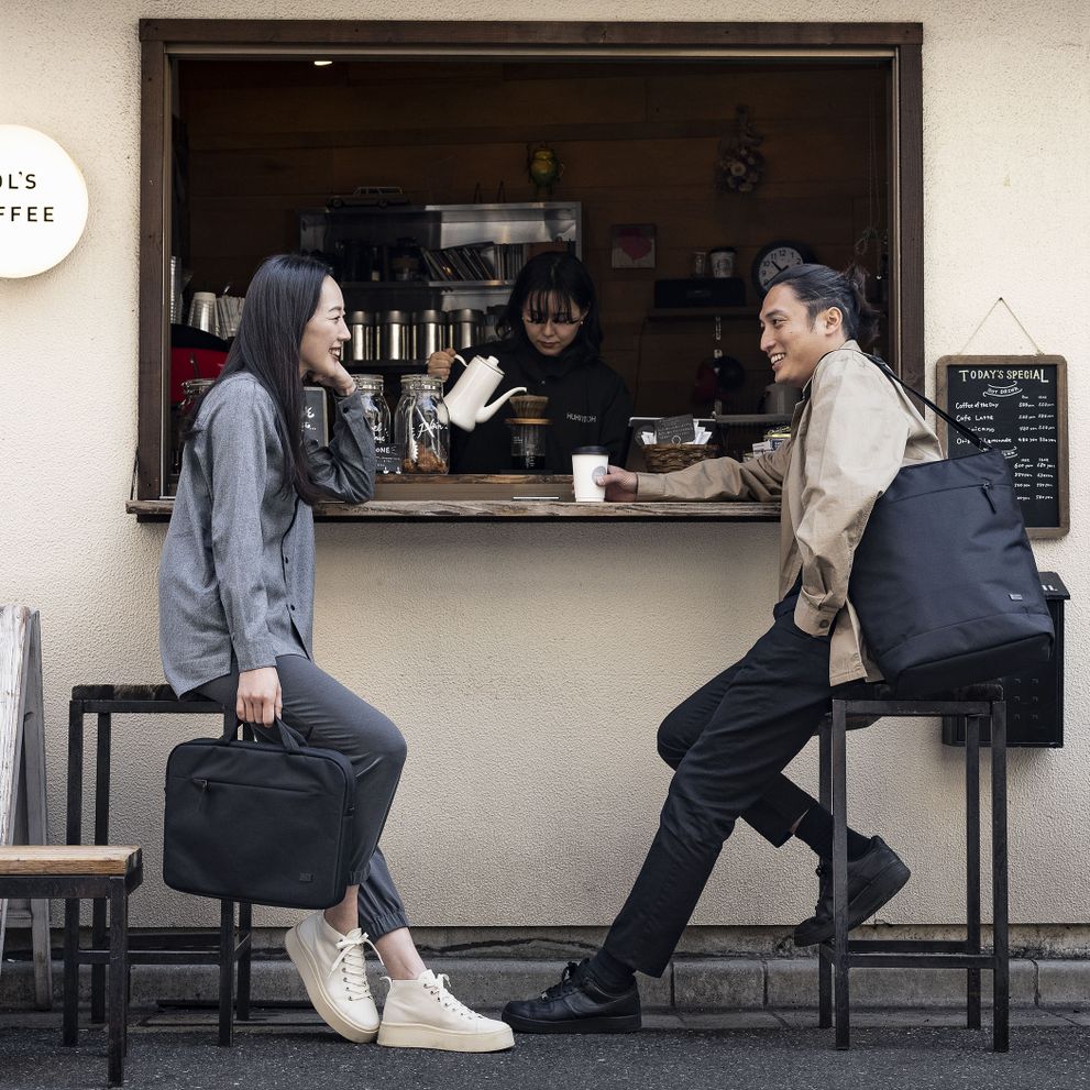A man and woman drink a coffee at a coffee shop while holding the Case Logic Invigo briefcase and tote.