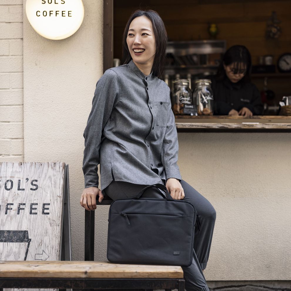 A woman stands outside of a coffee shop laughing, holding a black Case Logic Invigo briefcase.