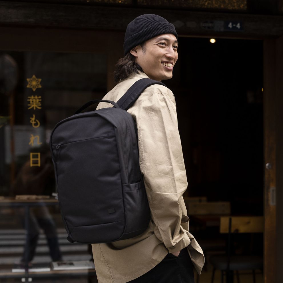 A man smiles wearing a black beanie and carrying a black Case Logic Invigo backpack.