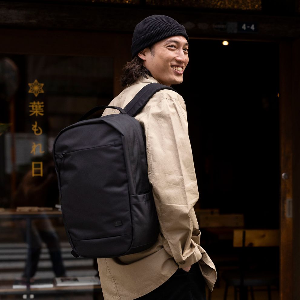 A man smiles wearing a black beanie and carrying a black Case Logic Invigo backpack.