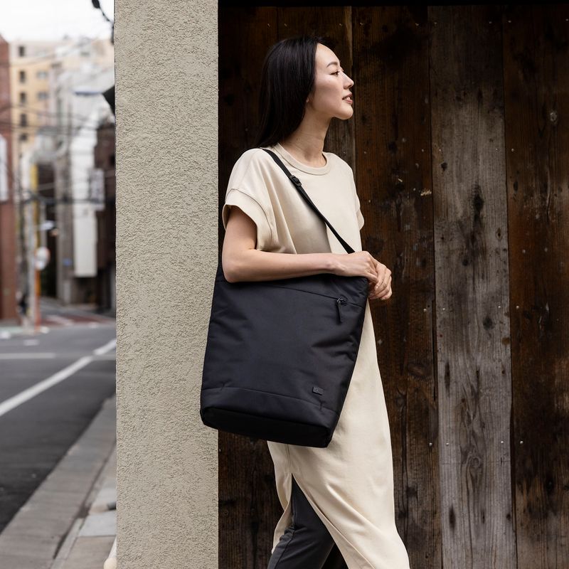 A woman leans on the wall by a city street, carrying a black Case Logic Invigo tote.