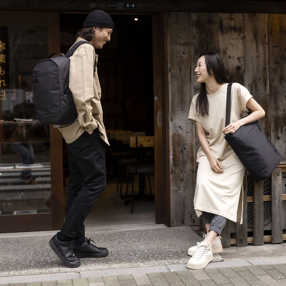 A couple laugh outside of a shop, holding black Case Logic Invigo tote and Case Logic Invigo backpack.