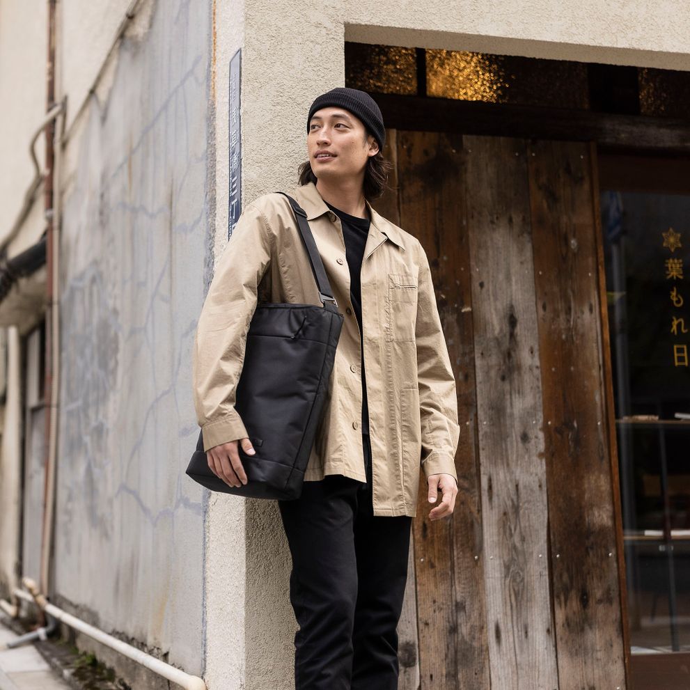 A man leans against a wall on a city street and carries a black Case Logic Invigo tote bag.