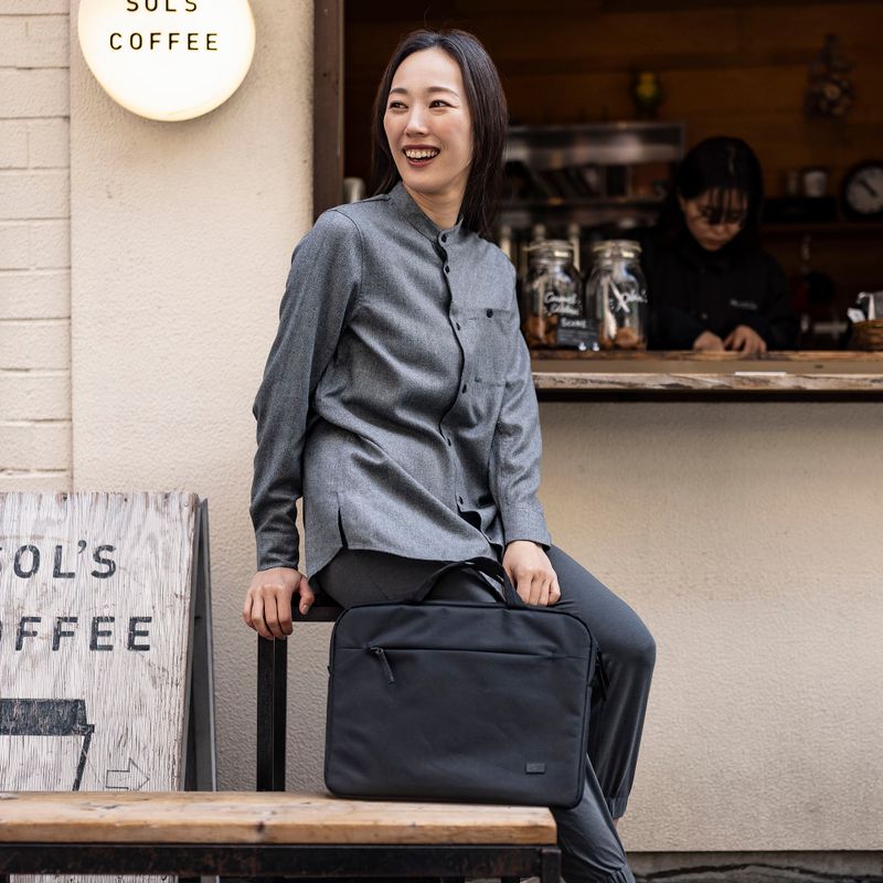 A woman stands outside of a coffee shop laughing, holding a black Case Logic Invigo briefcase.
