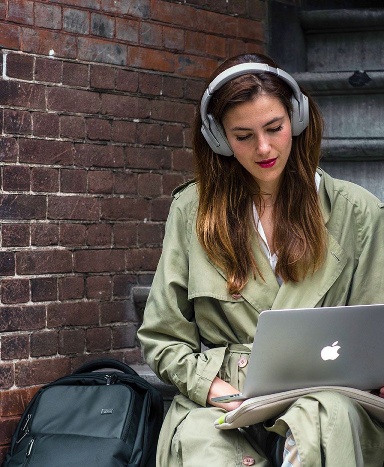 A woman sits on some steps with headphones, a laptop, and a Case Logic laptop backpack by her side.