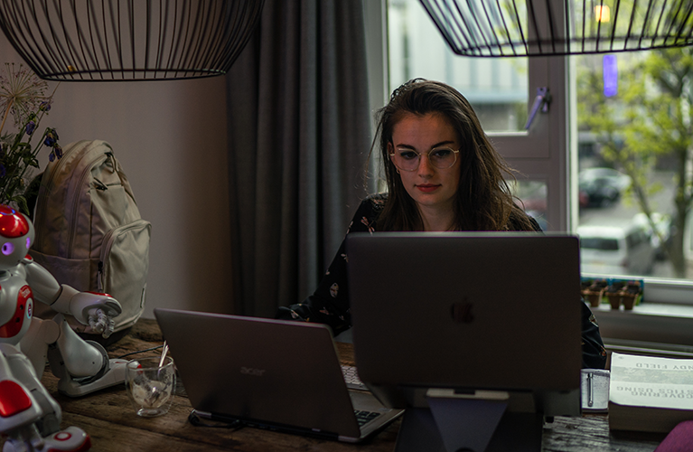 A students sits at a desk with her laptop, a small robot, and a Case Logic backpack.