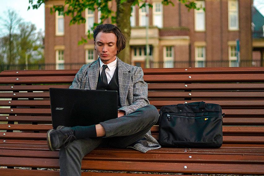 A man sits on a bench outside using his laptop with a Case Logic laptop briefcase beside him.