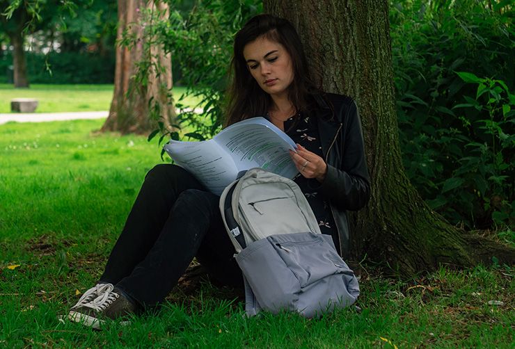 A woman sits against a tree looking through her notes with a Case Logic backpack for students beside her.