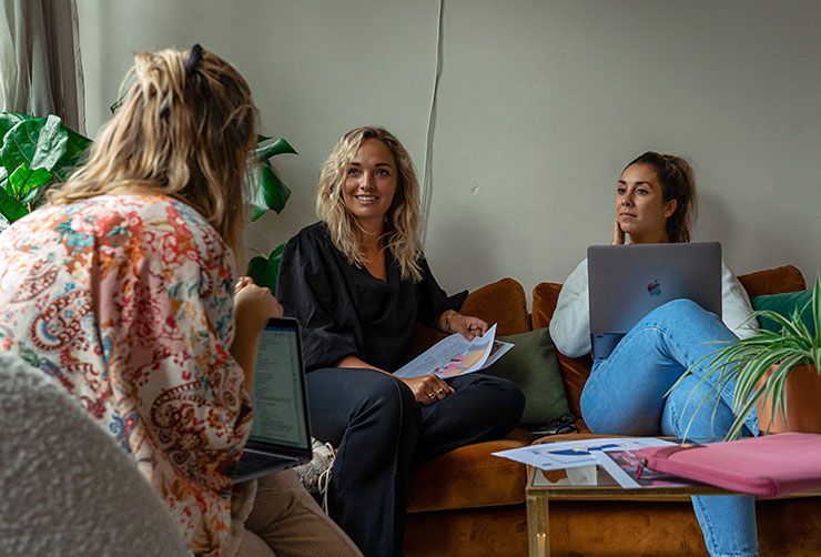 Three women chat around a table with laptops and a pink Case Logic laptop sleeve sits on the table.