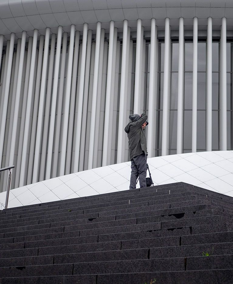 A man takes a photo of a white building with modern architecture, he is holding a Case Logic camera bag.