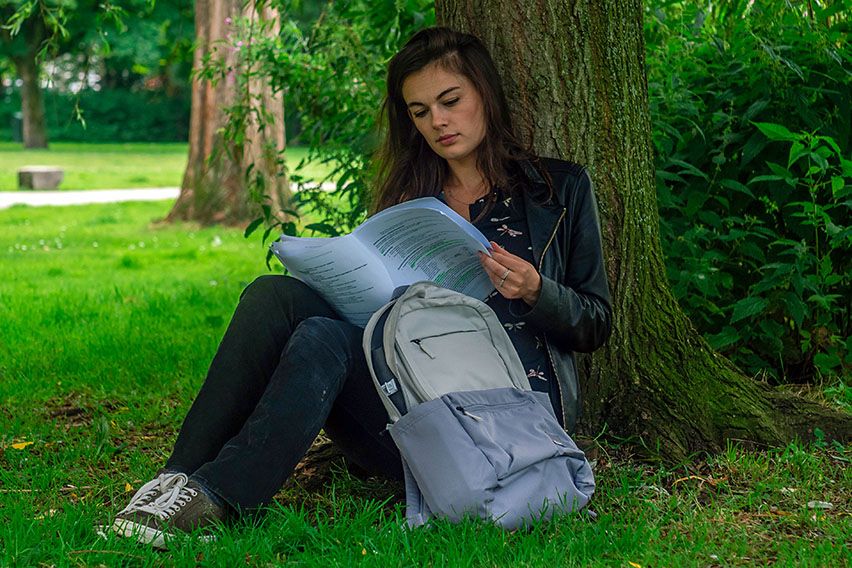 A woman sits against a tree looking through her notes with a Case Logic backpack for students beside her.