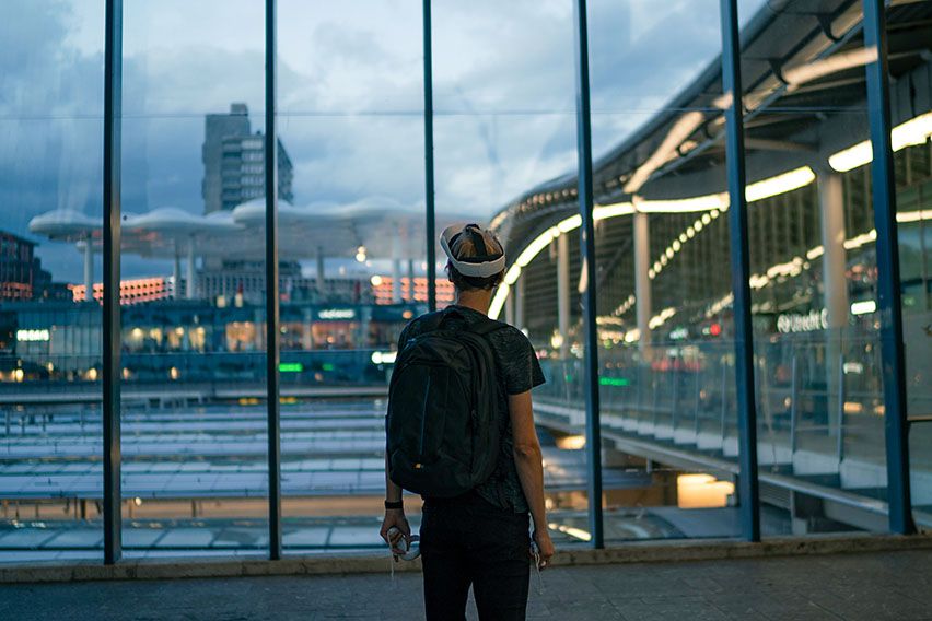 A man stands in a modern train station looking out a massive window with a VR set on and a Case Logic backpack.