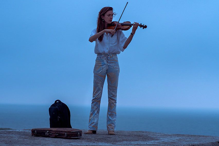 A woman stands beside a foggy seaside playing the violin with a Case Logic backpack beside her.