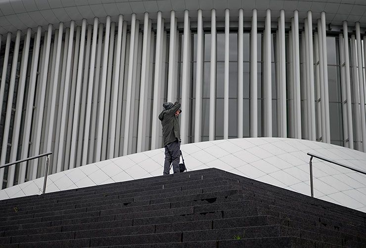 A man takes a photo of a white building with modern architecture, he is holding a Case Logic camera bag.
