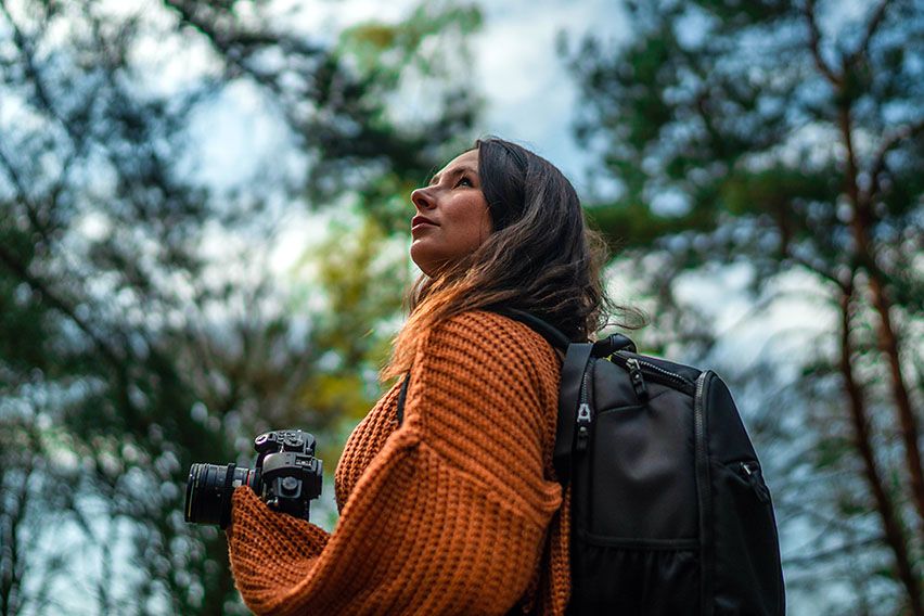 A woman in the forest looks up at the sky holding a camera and carrying Case Logic camera backpack.