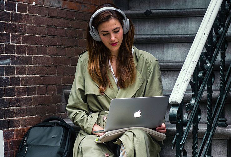 A woman sits on some steps with headphones, a laptop, and a Case Logic laptop backpack by her side.