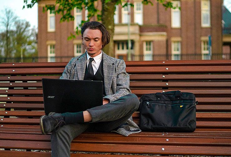 A man sits on a bench outside using his laptop with a Case Logic laptop briefcase beside him.