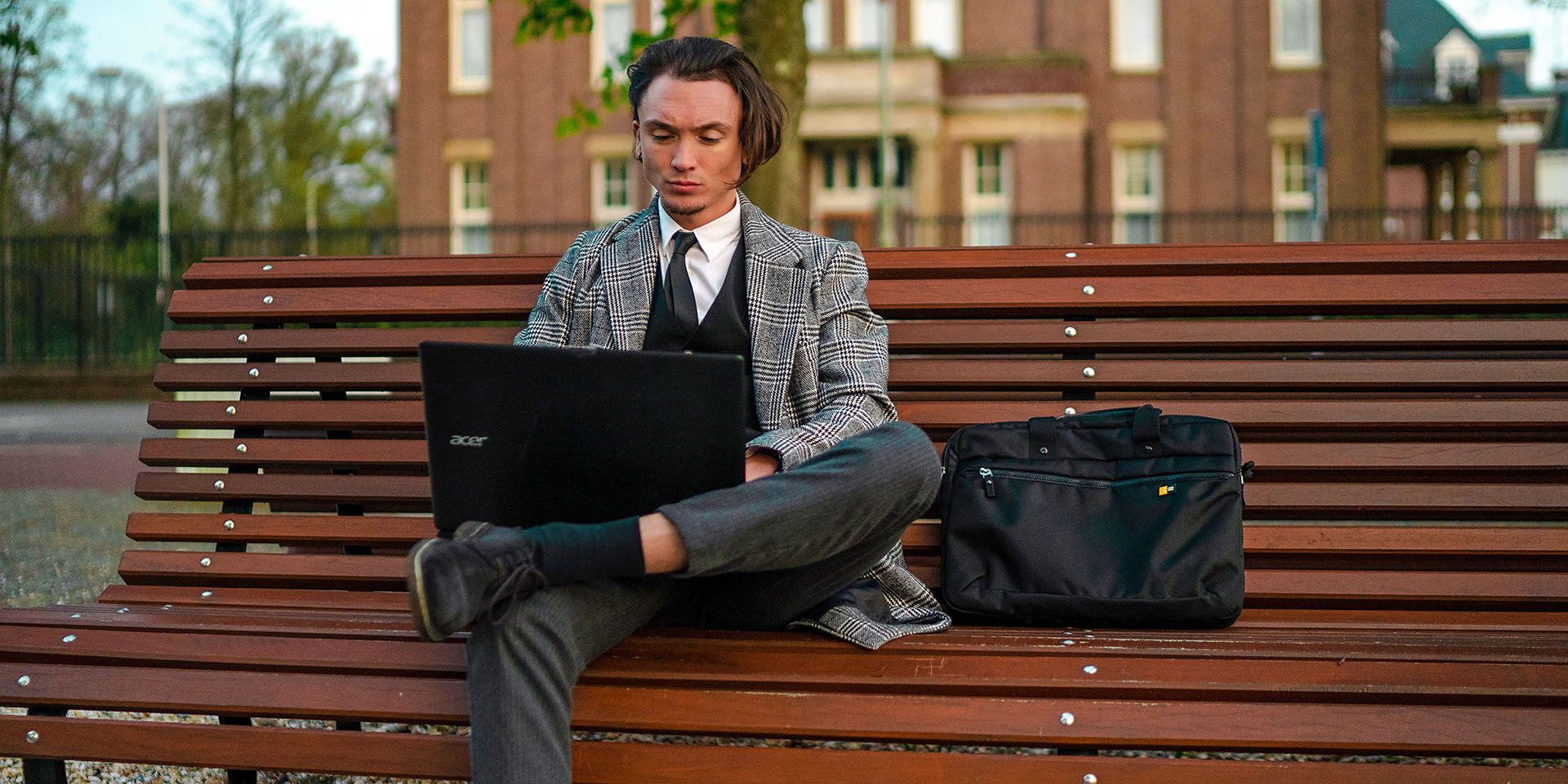 A man sits on a bench outside using his laptop with a Case Logic laptop briefcase beside him.